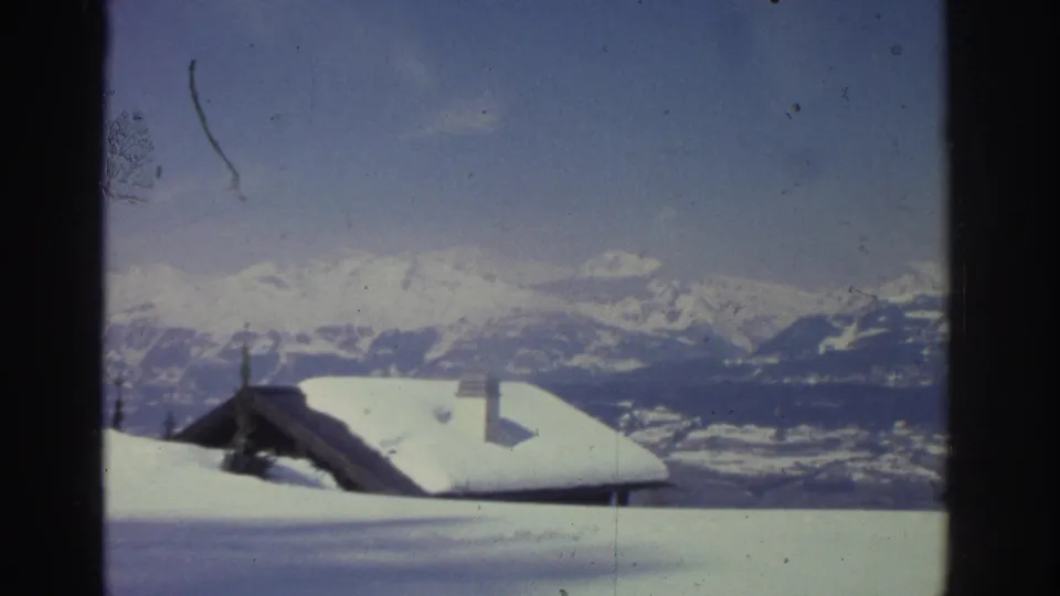 Closer framing of a snow-covered wooden chalet with chimney and distant mountain backdrop, Swiss Alps 1976, with a visible 8mm emulsion scratch at left.