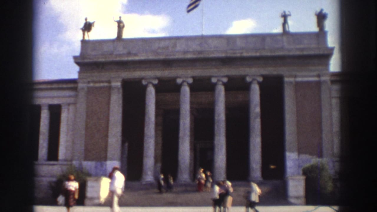 1976 Greece — wide 8mm view of a large neoclassical public building with classical columns, rooftop statues, a Greek flag, and 1970s pedestrians on the forecourt.