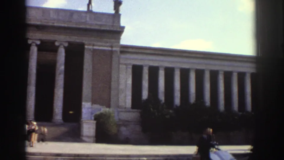 1976 Greece — lower-angle 8mm frame of a neoclassical colonnade; an overexposed light-leak streak dominates the upper centre; a few pedestrians and shrubs visible at the building base.