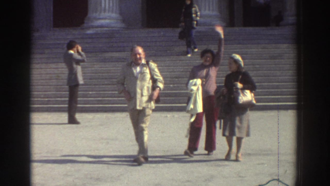 Three tourists descend wide stone steps before a building with large classical columns; one woman raises her hand in a wave toward the camera.