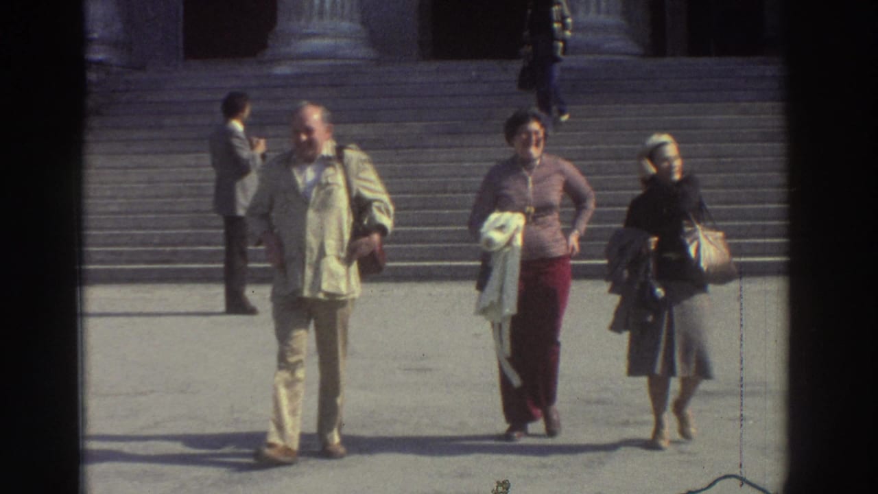 Three tourists pause on wide stone steps in front of large classical columns, facing the camera; a fourth visitor is visible in the background.