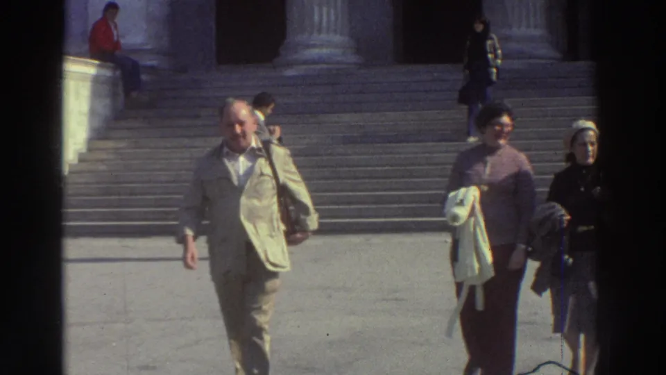 Tourists approach the camera on the steps of a neoclassical building in Greece, c. 1976; a seated figure in red and additional visitors appear on the steps behind them.