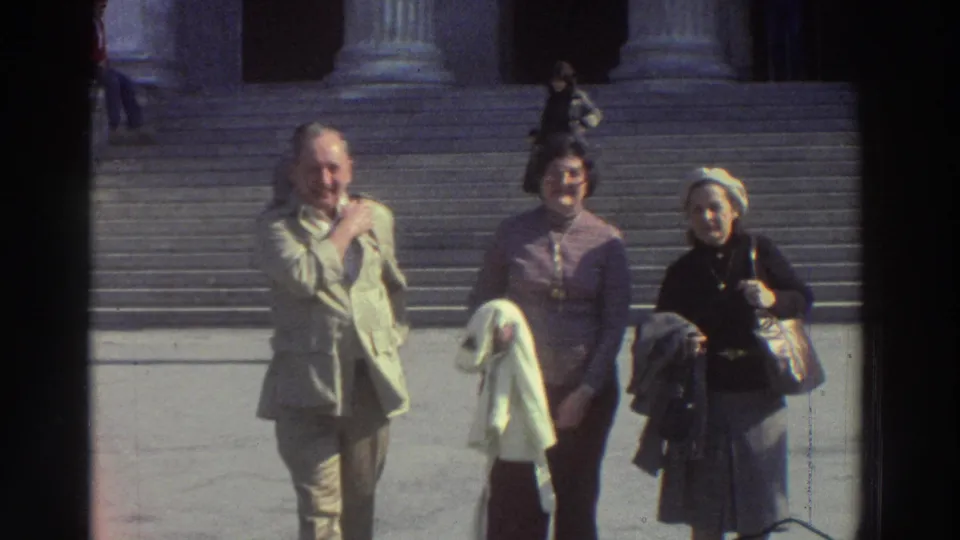 Close-up of three tourists at a neoclassical landmark in Greece, c. 1976; adult faces are clearly identifiable in this 8mm frame showing visible grain and edge vignette.