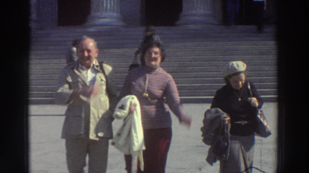 Man in light trench coat and two women tourists on wide stone steps at a neoclassical building, Greece, 1976; pink ribbed turtleneck and white beret visible.
