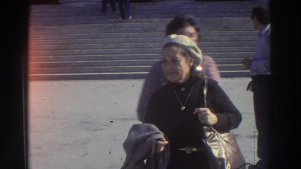 Close-up of woman tourist in dark jacket and white beret on stone steps, Greece, 1976; orange film light-leak at left edge and pendant necklace visible.