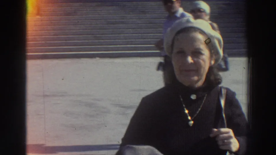 Smiling woman tourist in white beret and pendant necklace faces camera on stone steps, Greece, 1976; a second figure is visible behind her.