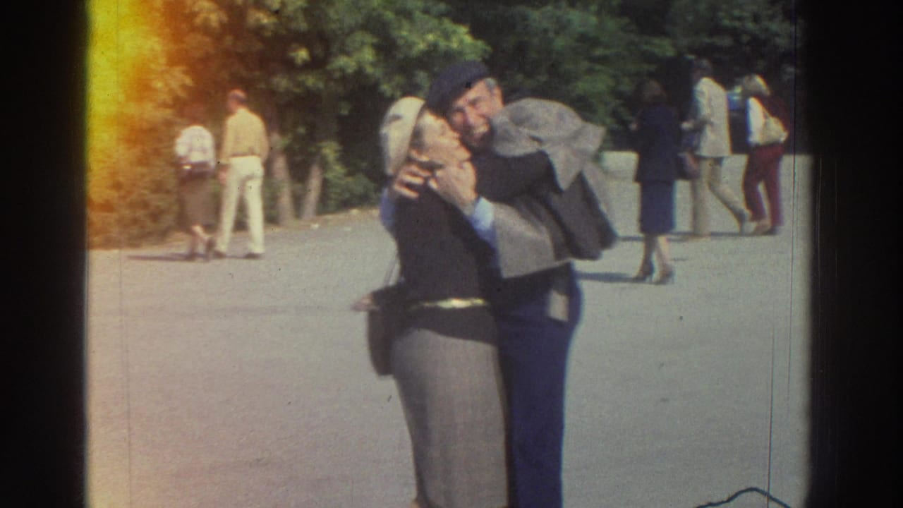 Elderly couple in 1970s dress embrace on a park walkway in what metadata describes as Greece, 1976; tree canopy and other pedestrians visible behind them.