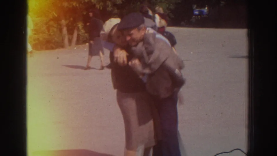 Close-up of an elderly couple embracing outdoors, consistent with Greece 1976 metadata; light wall behind them, trees at left, second figure partially visible at right edge.