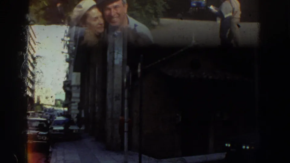 Elderly couple smiling on a narrow urban street consistent with Greece 1976 metadata; 1970s-era cars and multi-story buildings visible; film damage or exposure artifact at left edge.