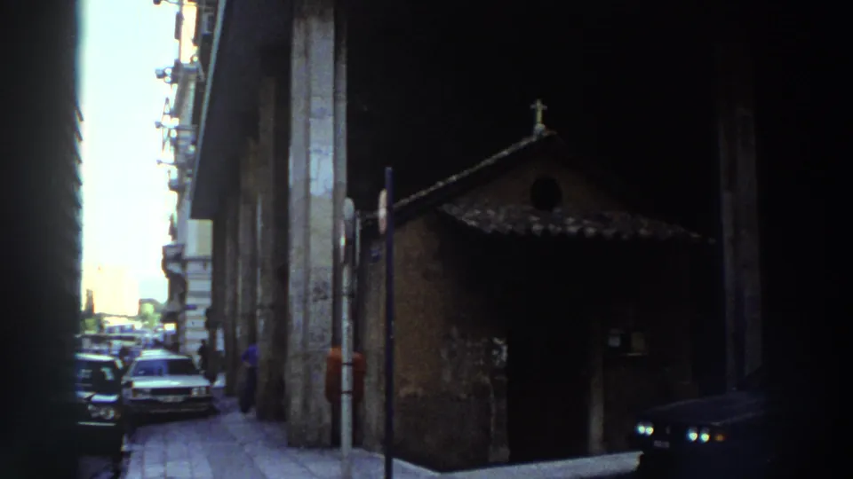 Narrow street in what metadata describes as Greece, c.1976; small stone or brick church with dark tiled roof at right, 1970s-era automobiles, multi-story buildings on both sides, clear sky above.