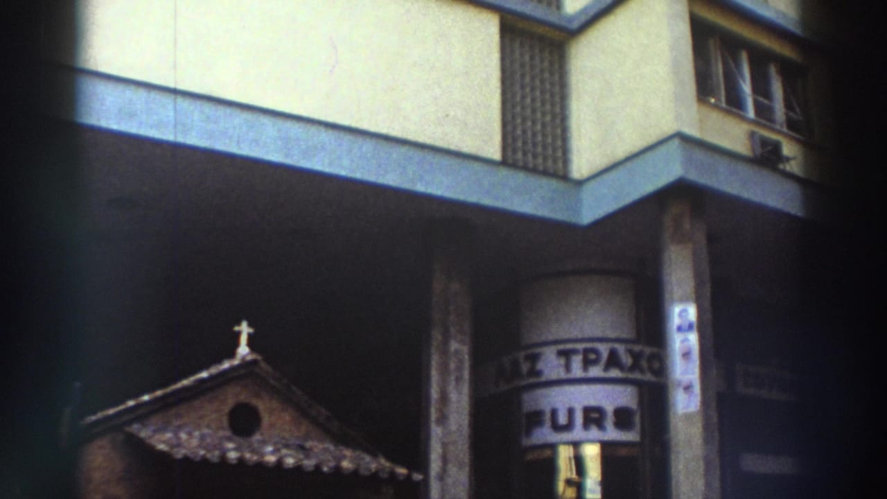8mm home-movie frame from Greece, circa 1976, showing a small Byzantine-style chapel beside a modern apartment building with partially visible Greek-language signage.