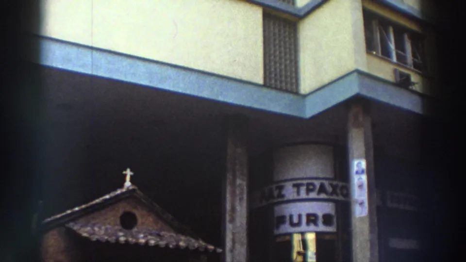 8mm home-movie frame from Greece, circa 1976, showing a small Byzantine-style chapel beside a modern apartment building with partially visible Greek-language signage.