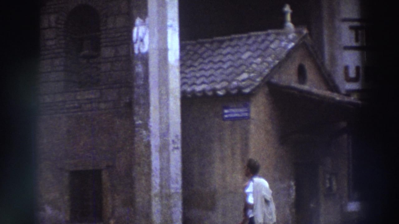 Man in light shirt walks past a small stone chapel with a tiled roof and cross; blue street sign visible on chapel wall.