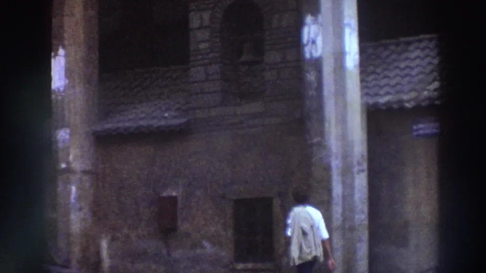 8mm home-movie frame from Greece, circa 1976, showing a man walking past a small stone chapel with tiled roof, seen from behind.