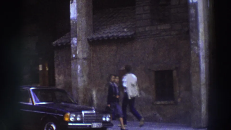 8mm footage from Greece, circa 1976, showing two pedestrians and a dark classic Mercedes sedan parked beside a small stone chapel on a narrow town street.