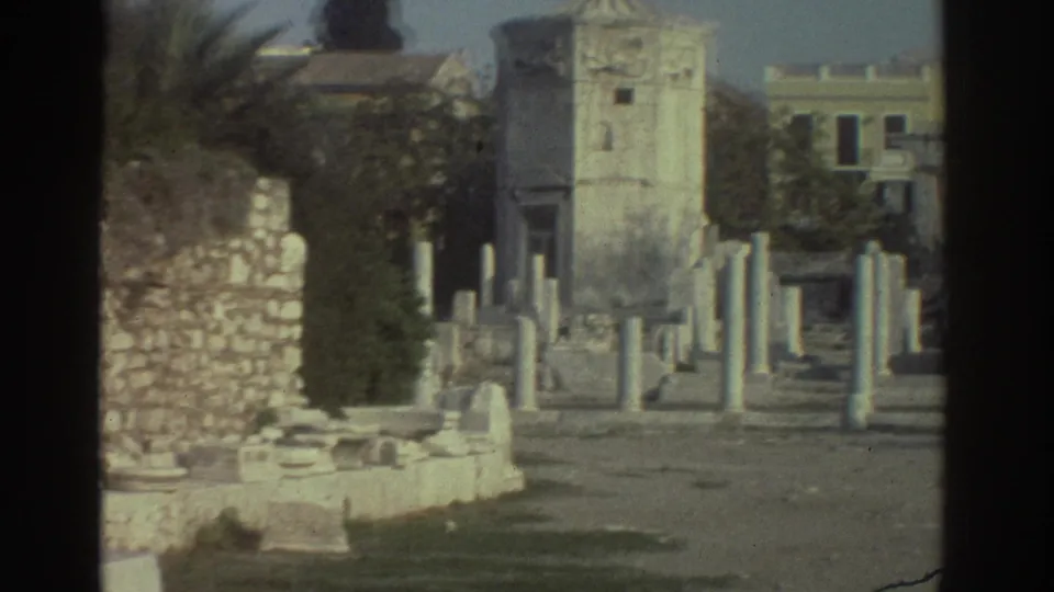 8mm tourist frame from Greece, circa 1976, showing rows of column remnants, a stone tower structure, and mid-century modern buildings visible behind the ruins.
