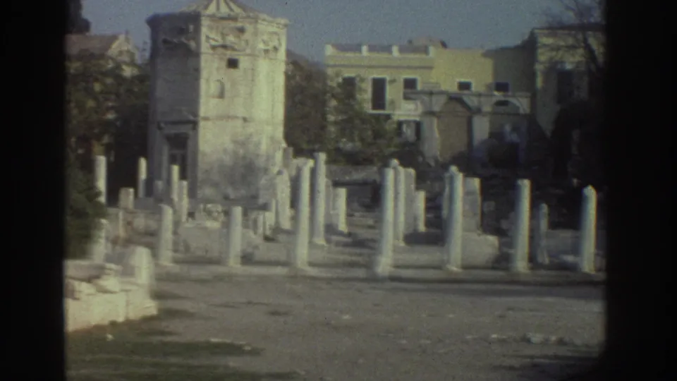 8mm pan frame from Greece, circa 1976, showing foreground column stumps, a ruined stone structure with carved relief decoration, and modern urban buildings beyond.