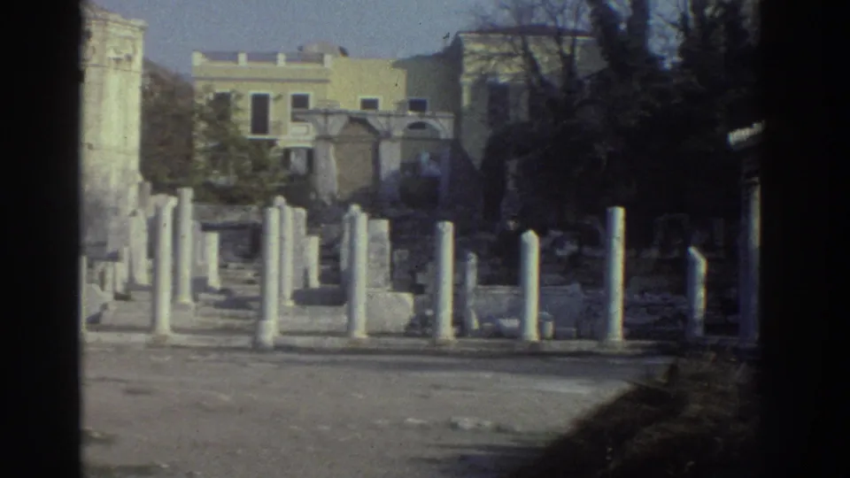 Closing frame of an 8mm tourist pan across Greek archaeological ruins, circa 1976, showing column rows, a carved stone monument, and an urban backdrop.