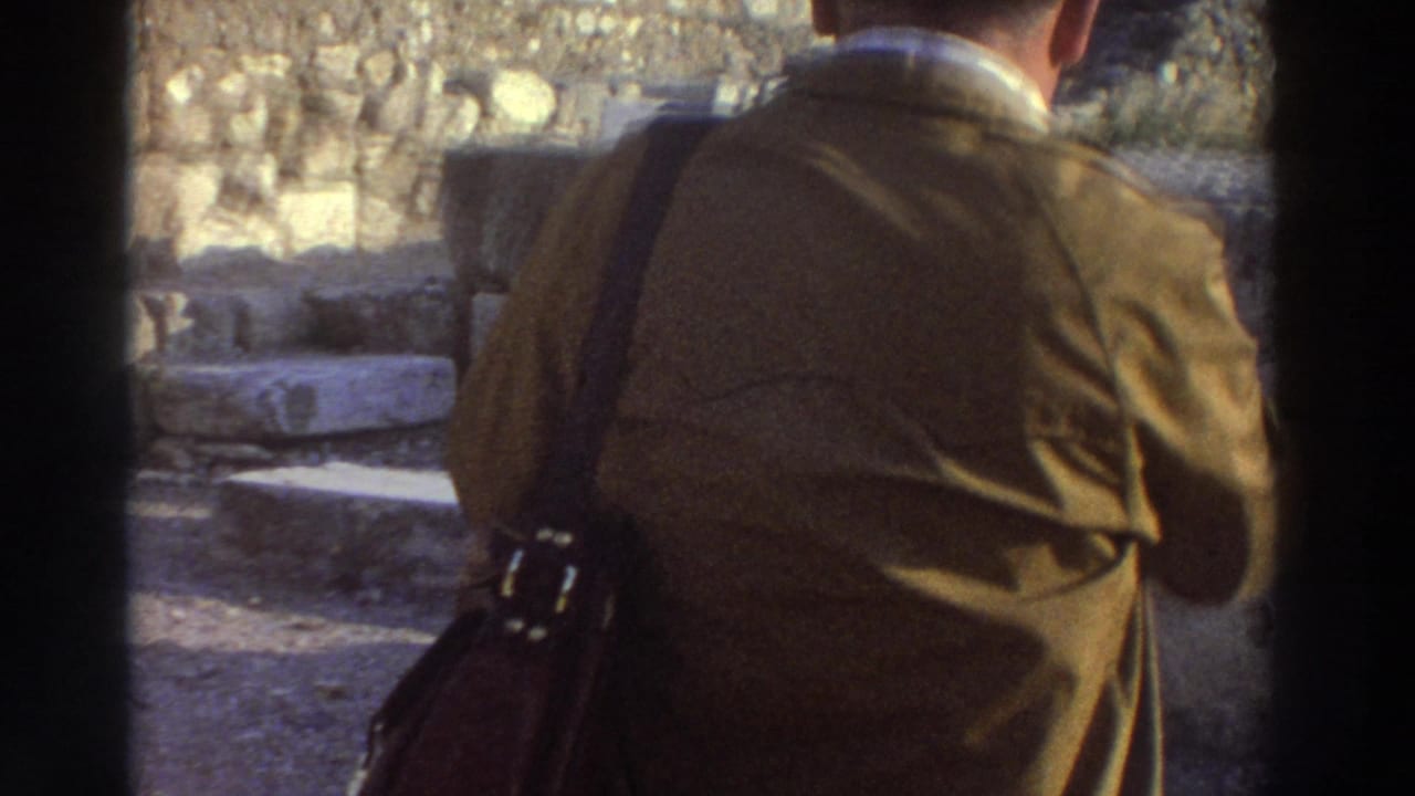 Close-up of a man's back and shoulder in a brown jacket with a leather shoulder bag; stone ruins visible behind him.