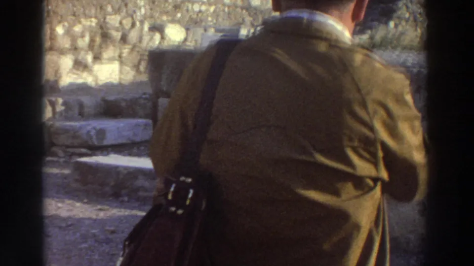Rear close-up of a man in a brown jacket carrying a leather shoulder bag at a Greek archaeological site, circa 1976 8mm footage.