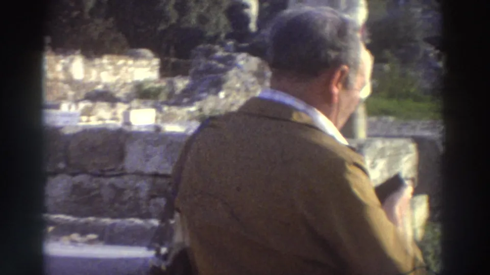 Man in 1970s brown jacket seen in partial profile apparently holding a camera, stone ruins in background — 8mm home movie, Greece, 1976.