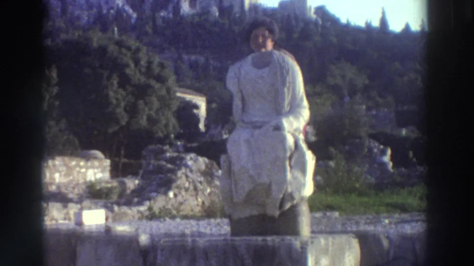 Robed classical statue on a stone plinth at Greek archaeological ruins, circa 1976; green hillside with a possible distant structure visible behind.