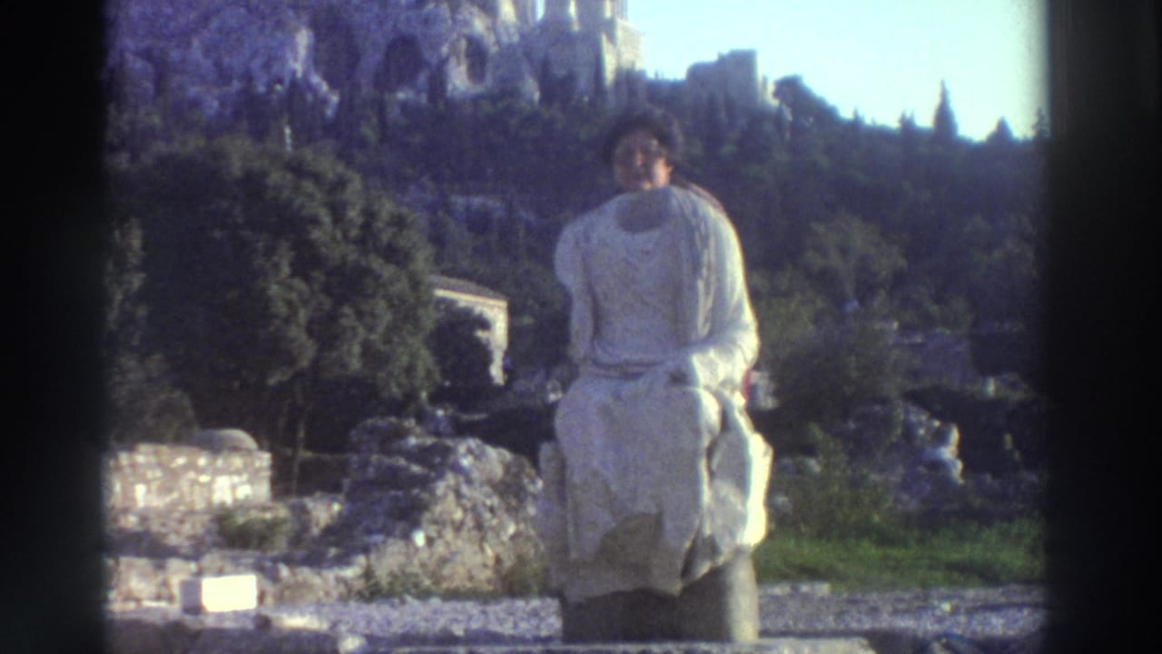 Woman with dark curly hair and large-frame glasses in a white toga-like garment poses at a Greek archaeological site, circa 1976, with ancient hillside ruins behind her.