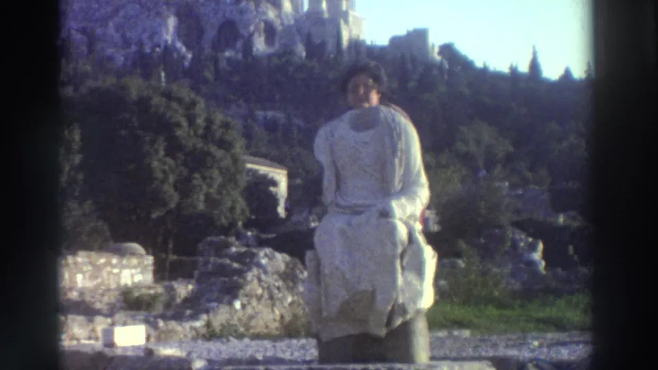 Woman with dark curly hair and large-frame glasses in a white toga-like garment poses at a Greek archaeological site, circa 1976, with ancient hillside ruins behind her.