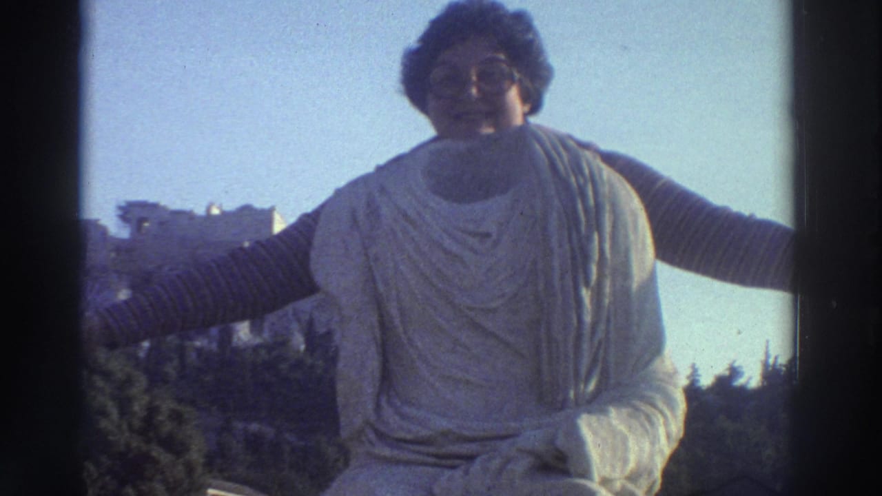 Close-up of woman with glasses and curly hair extending arms outward in a draped white garment, ancient hilltop structure visible in background under blue sky.