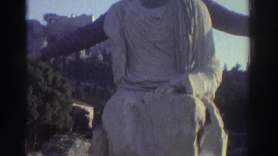 The trick revealed: a headless draped stone statue torso occupies the foreground while the woman's striped-sleeve arm extends from behind it, at a Greek archaeological site circa 1976.