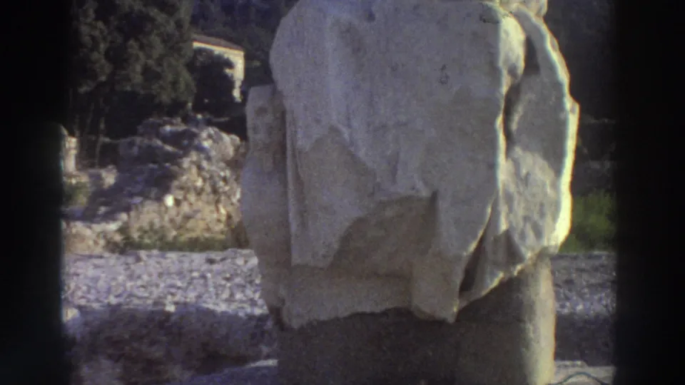 Stone statue torso fragment on pedestal at a Greek archaeological site circa 1976, woman's arm still extended from behind it, ancient ruins and gravel scattered in background.