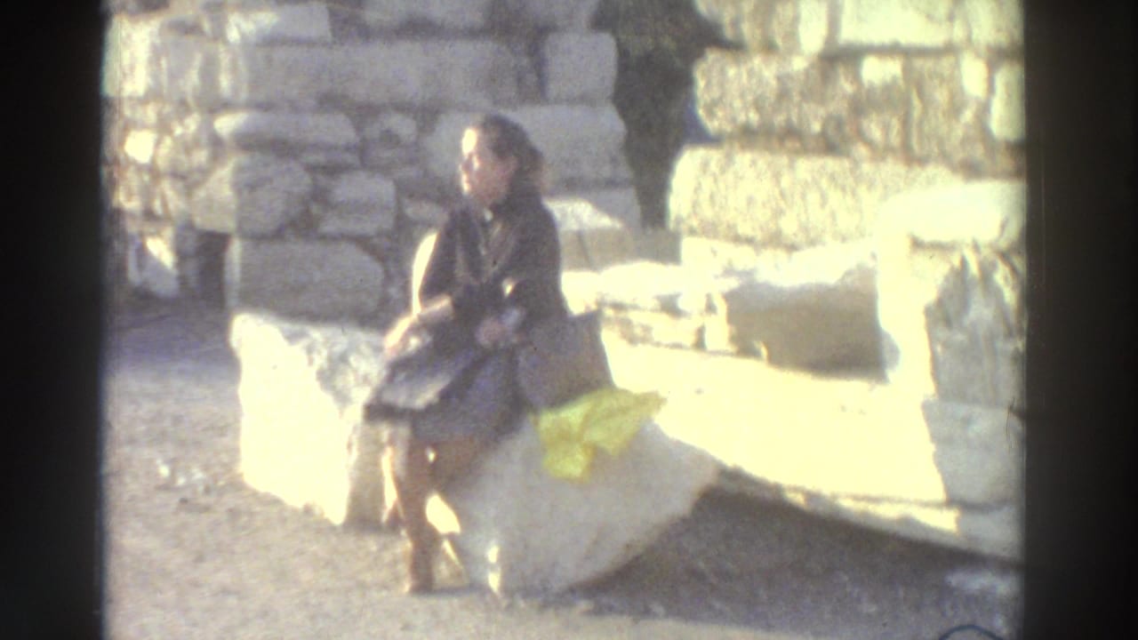 Young woman in dark coat sits on ancient stone ruins, looking away from camera; yellow cloth beside her on the stone.