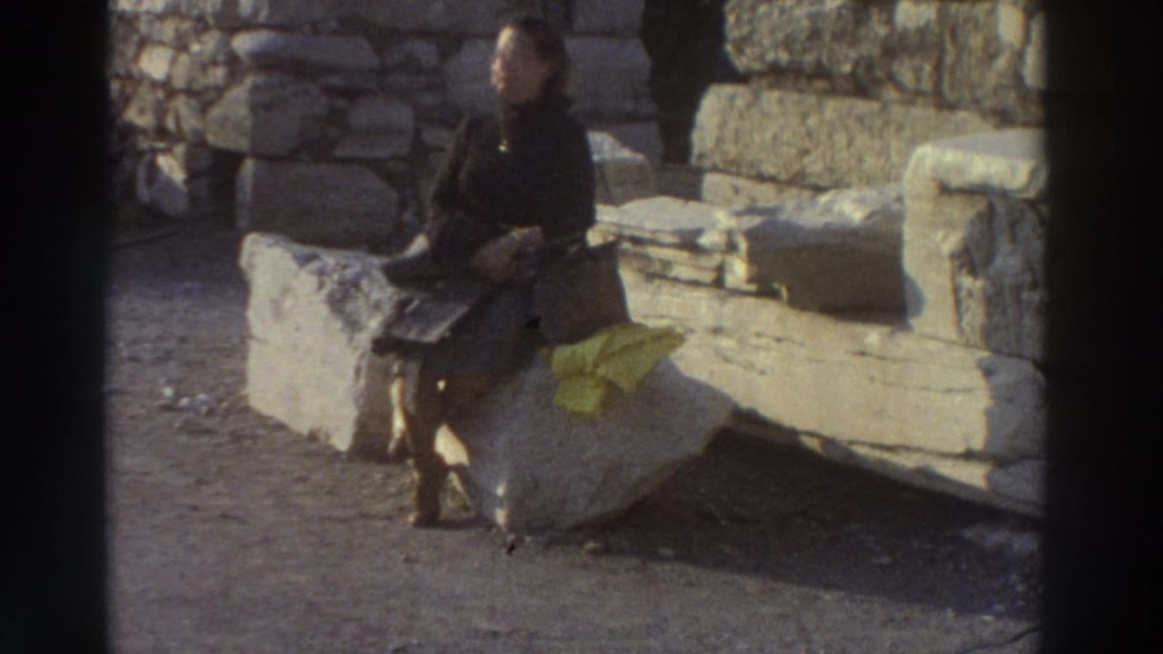 Young woman in dark coat seated on a flat stone block looking toward camera; yellow cloth on stone beside her, ancient wall behind.
