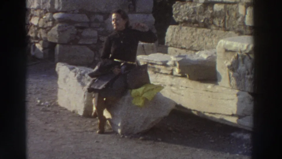 Young woman smiling as she turns toward the camera while seated on ancient stone blocks at a Greek site, circa 1976; yellow cloth visible beside her.