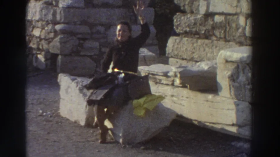 Young woman waving and smiling at the camera while seated on ancient stone ruins in Greece, circa 1976; another person partially visible at right edge.