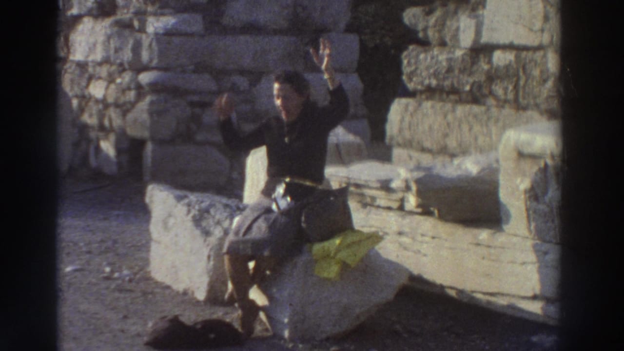 Woman tourist seated on large cut-stone ruins in Greece, arms raised in celebration, 8mm home movie circa 1976