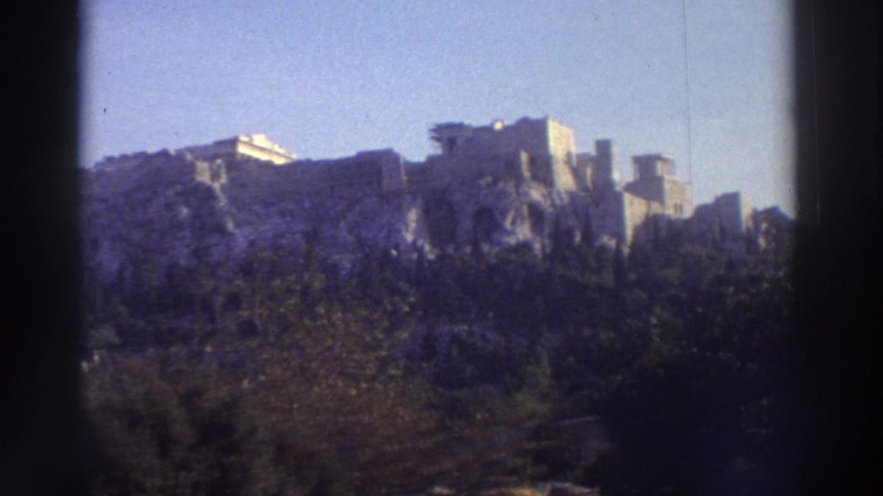 8mm shot of ruined marble structures atop a rocky hill, framed by trees and a clear sky.