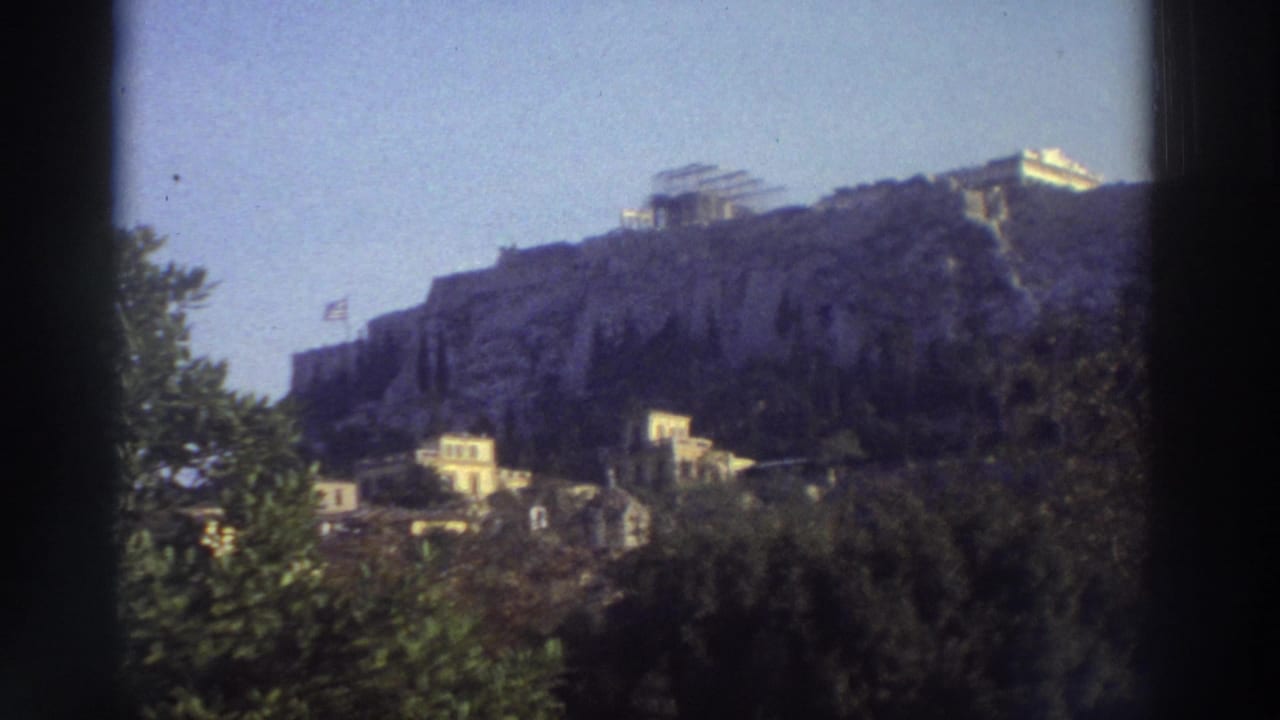 Acropolis of Athens viewed from below, rocky hill with ruins on summit, Greek flag on slope, trees in foreground.