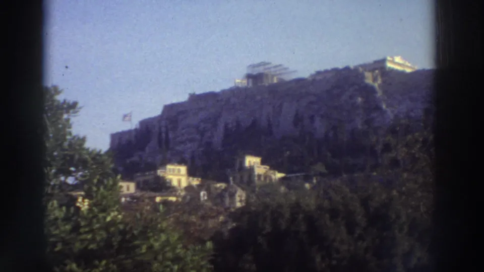 Distant view of the Acropolis of Athens from ground level, c. 1976, with Greek flag on the hillside and Plaka-area buildings at the base.