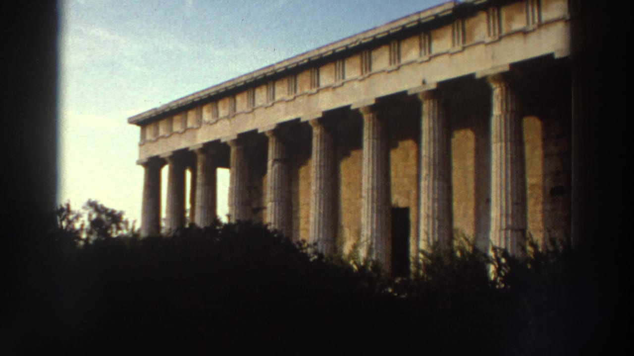 Frontal view of a Doric temple façade with colonnade and entablature, seen through foreground foliage against blue sky.