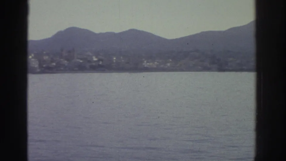 Wide view of a Greek coastal bay with a hillside settlement and mountains in the distance, circa 1976, 8mm home movie with slight haze.