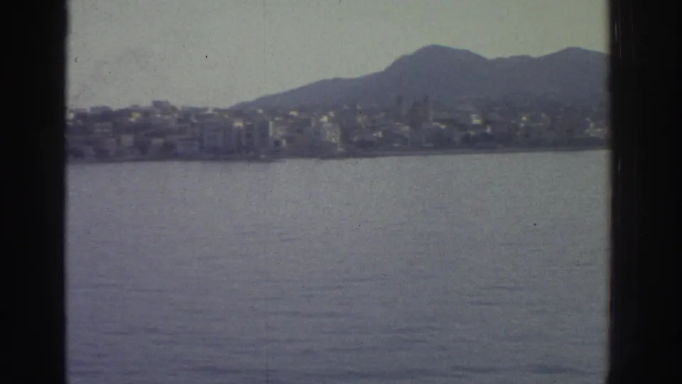 Continued pan over a Greek bay toward a coastal settlement beneath a mountain ridgeline, circa 1976, 8mm film.