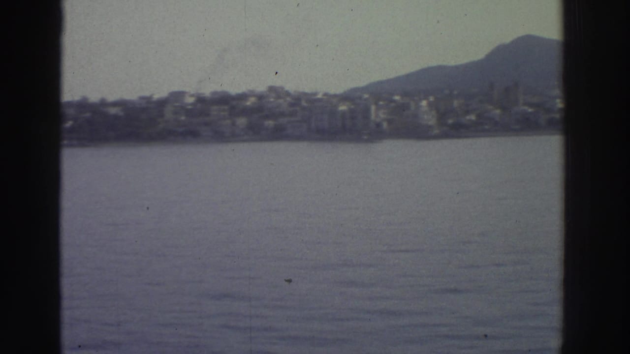 Greek coastal town on rolling hills seen across calm water from a boat, 8mm film with dark frame borders.