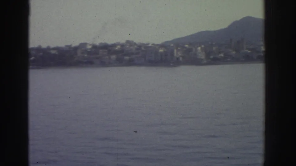 1976 Greece — white buildings spread across a hillside above calm blue-grey water, viewed from a moving vessel; 8mm home movie with grain and dark borders.