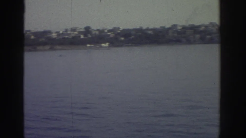 1976 Greece — continued pan across a hillside settlement of white and light-coloured buildings above a calm Mediterranean shoreline; 8mm home movie.