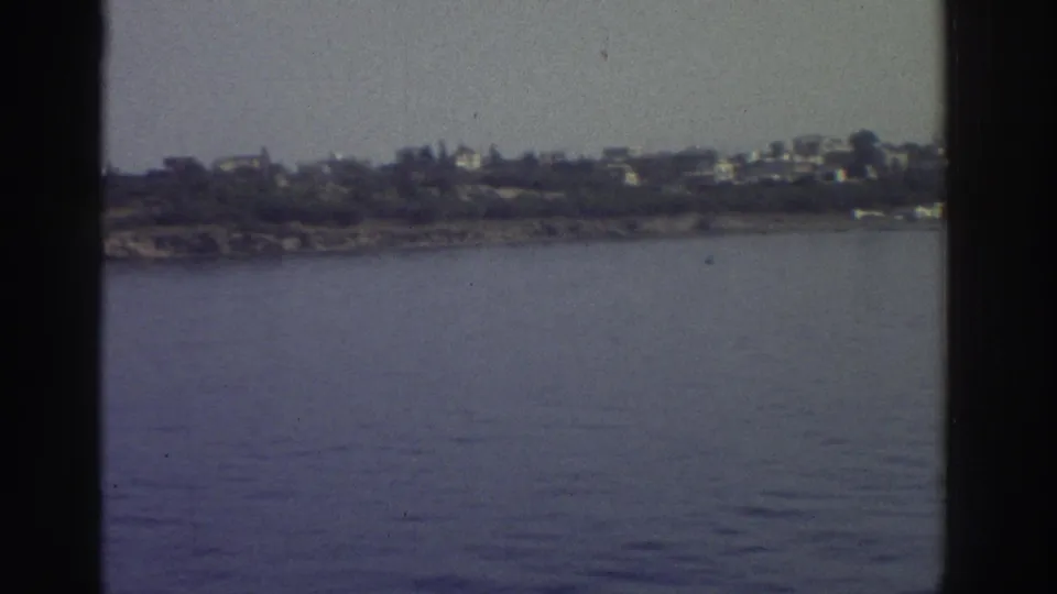 1976 Greece — low rocky headland with sparse white buildings and coastal vegetation at the waterline, viewed from a moving vessel; 8mm home movie.