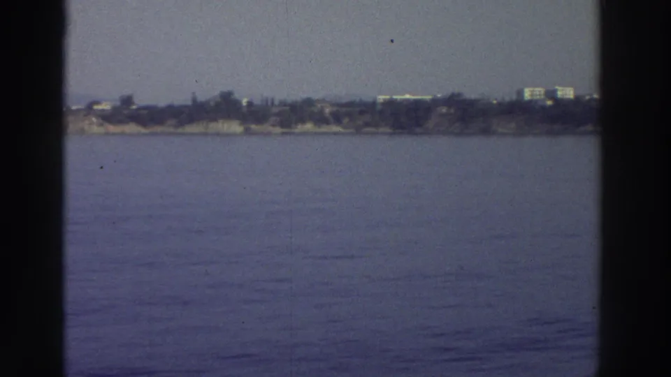 1976 Greece — rocky coastal point with sparse buildings and shrubs at the waterline, end of the panning sequence; 8mm home movie.