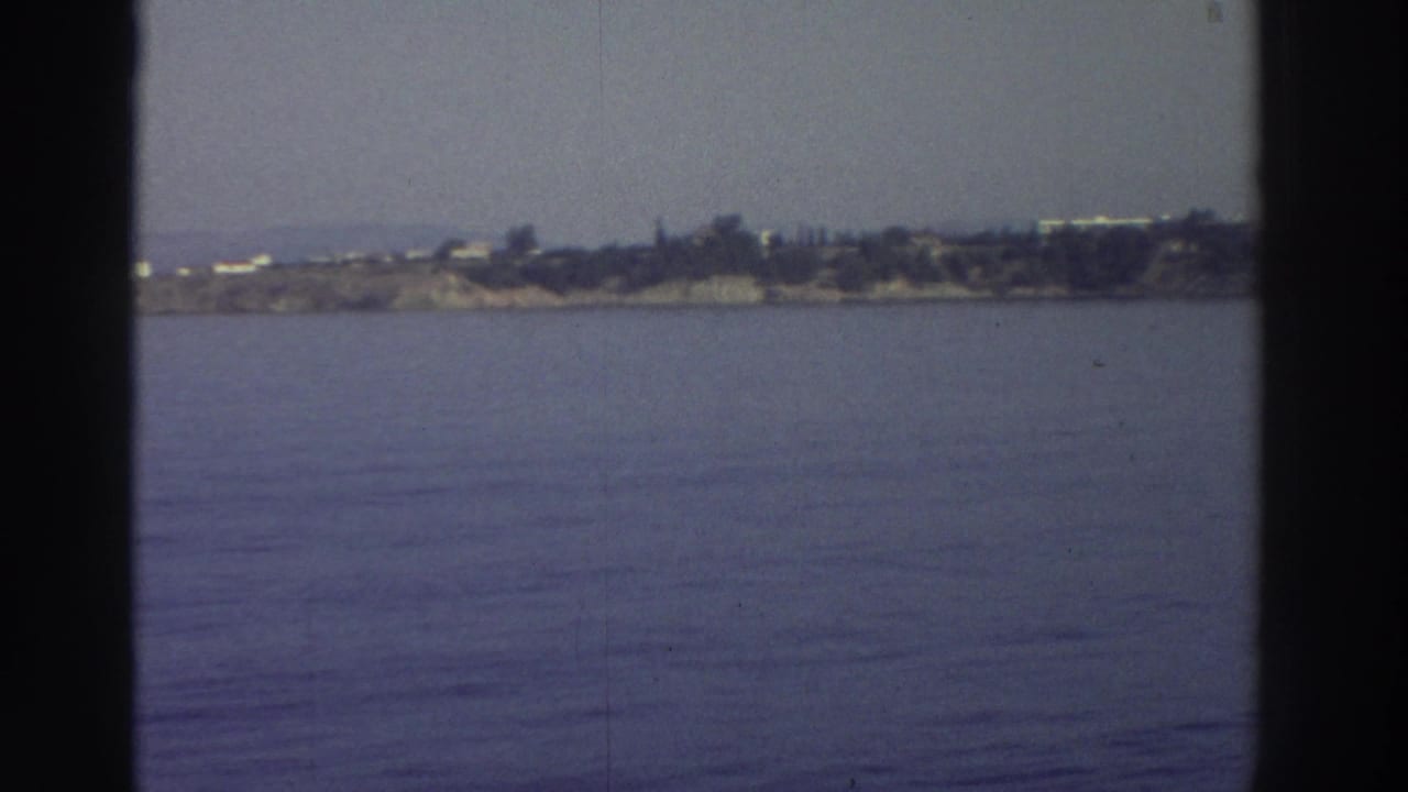 8mm home-movie frame from a moving vessel, c.1976 Greece — calm Aegean water and a low rocky coastline with white buildings visible in the distance.