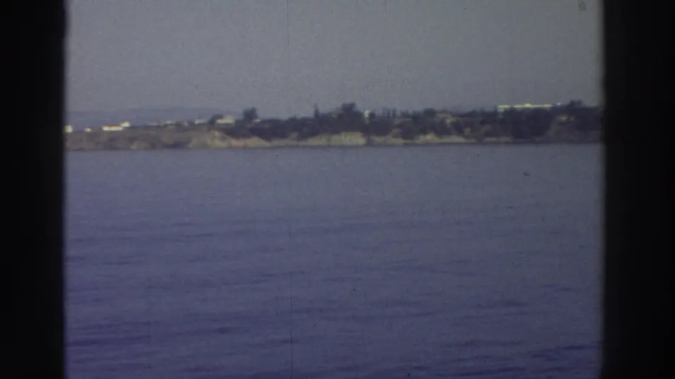 8mm home-movie frame from a moving vessel, c.1976 Greece — calm Aegean water and a low rocky coastline with white buildings visible in the distance.
