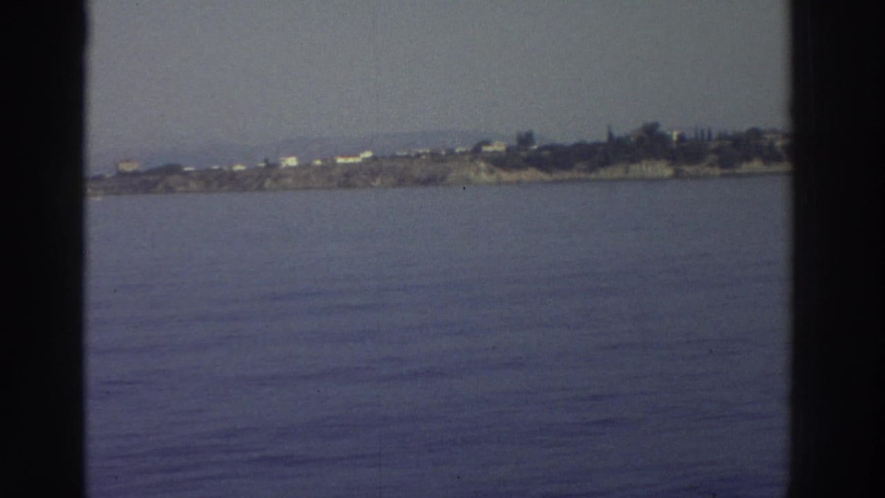 Calm blue-purple water with a rocky Greek headland and white buildings visible in the distance, seen through a ship's window.